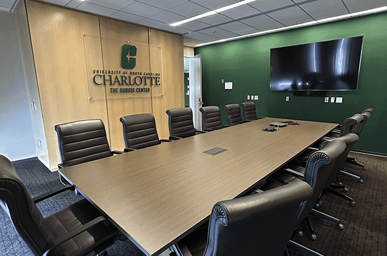 a large boardroom with a conference table surrounded by executive chairs, the UNC Charlotte logo hangs on a handsome wood wall